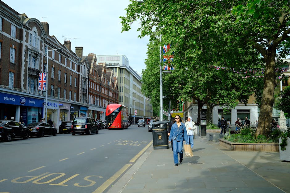 A busy street scene in St John's Wood captures two women walking along the pavement, one wearing a blue denim jacket and sunglasses, the other with a white headscarf and beige handbag, with vehicles i