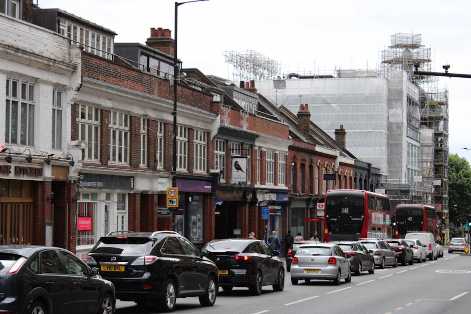 A busy street scene in St John's Wood captures two women walking along the pavement, one wearing a blue denim jacket and sunglasses, the other with a white headscarf and beige handbag, with vehicles i