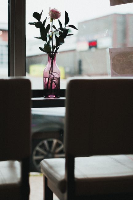 Inside a room with a large window, a narrow black table holds a pink glass vase containing a single pink flower with green leaves, positioned centrally on the table. The window behind the vase shows an outdoor view of a parking area with blurred cars and buildings. In the foreground, two partially visible brown upholstered chairs face the window, suggesting a dining or sitting area. The scene is lit by natural daylight, creating a calm atmosphere relevant to home relocation or moving preparations. The setting reflects the interior environment of a residential or commercial property before transportation, aligning with the services offered by St John's Wood Movers for furniture transport, packing, and moving logistics within house removals.