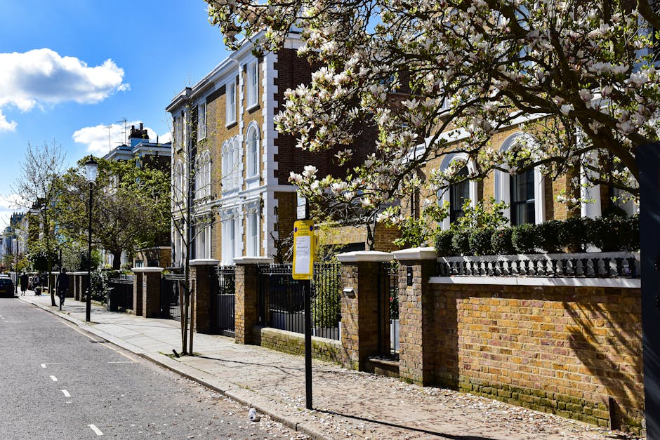A residential street scene in St John's Wood showing a row of Victorian-style houses with brick facades, white window frames, and decorative architectural details. The pavement is lined with young trees, some blossom in white flowers, and black metal lampposts stand along the sidewalk. A black metal fence with brick pillars surrounds the front yards of the houses, which are accessed via gates. The image captures part of a house’s front garden with neatly trimmed bushes and white window blinds visible through the windows. The sky above is partly cloudy with patches of blue, and the natural daylight highlights the textures of the brickwork, trees, and pavement. This setting reflects a typical London neighbourhood, where house removals or home relocation services by St John's Wood Movers may involve careful packing and loading of furniture, boxes, and appliances from within the property, with equipment such as trolleys or straps potentially seen during such activities outside the residence.